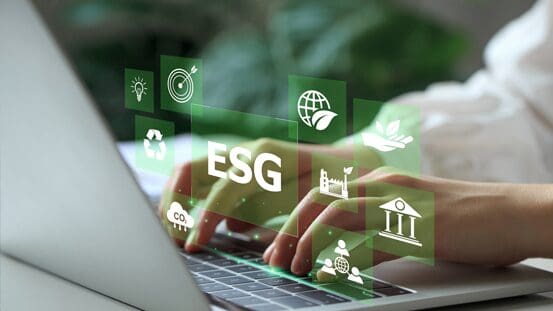 Close-up of hands typing on a laptop keyboard with virtual green icons representing ESG (Environmental, Social, Governance) concepts, such as recycling, energy, global, and institutional symbols, overlaying the image.