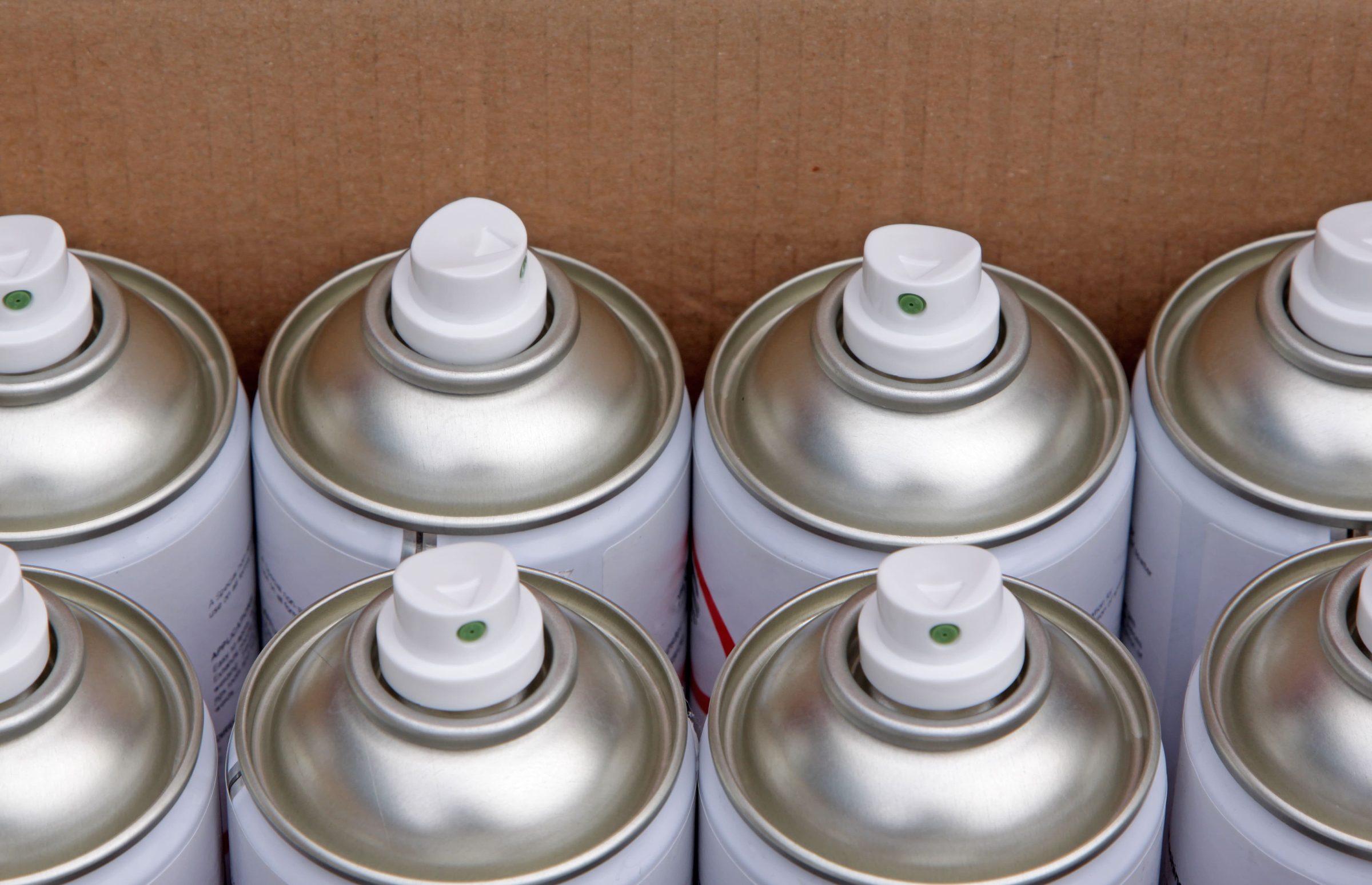Close-up view of several silver spray paint cans with white nozzles, arranged closely together inside a cardboard box.