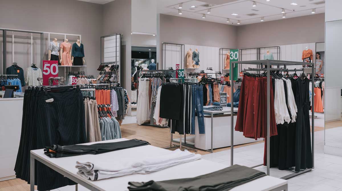 Clothing store interior with neatly organized racks and tables displaying folded and hanging clothes. Sale signs with discounts of 30% and 50% are visible around the store. The lighting is bright and the space is tidy.