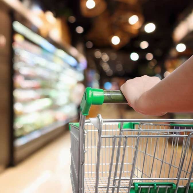 A person’s hand is pushing a shopping cart down an aisle in a brightly lit grocery store, with shelves of products visible in the blurred background.