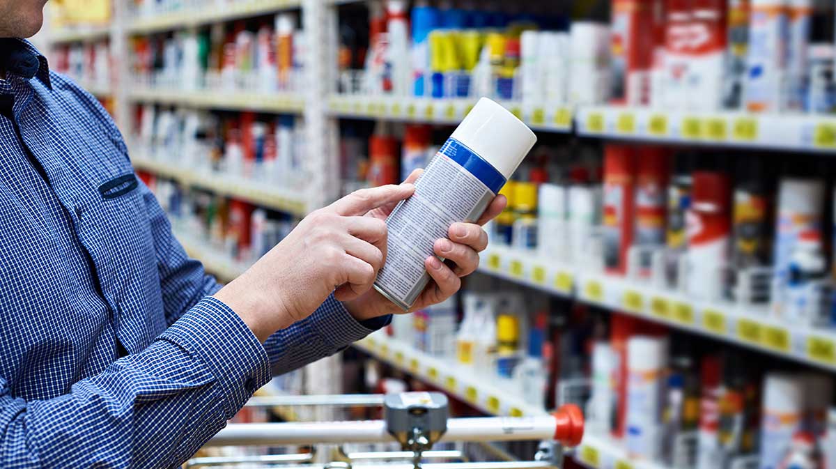 A person wearing a blue checkered shirt reads the label on a can in a store aisle stocked with similar spray cans, possibly in the cleaning or automotive section.
