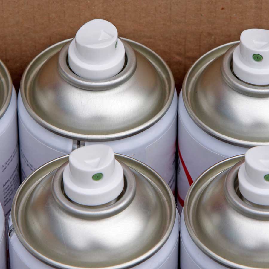 Close-up view of the tops of several aerosol spray cans arranged next to each other, showing white plastic nozzles and silver metal rims. The background appears to be a cardboard box.