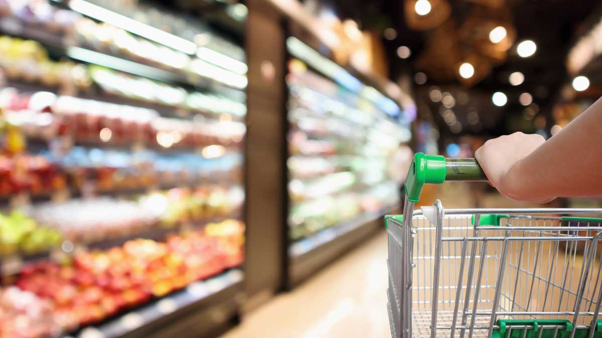 A person pushes a shopping cart down an aisle in a brightly lit grocery store, with shelves of colorful produce and products visible in the background.