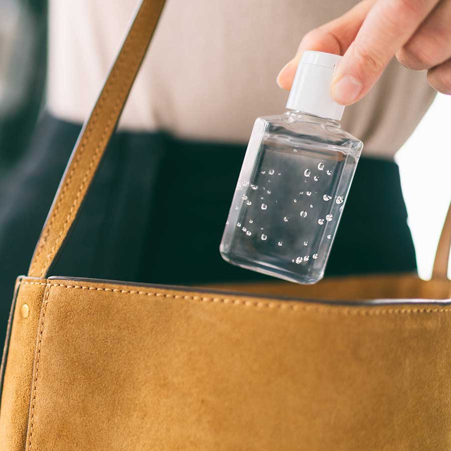 A person places a small clear bottle of hand sanitizer into a tan suede handbag.