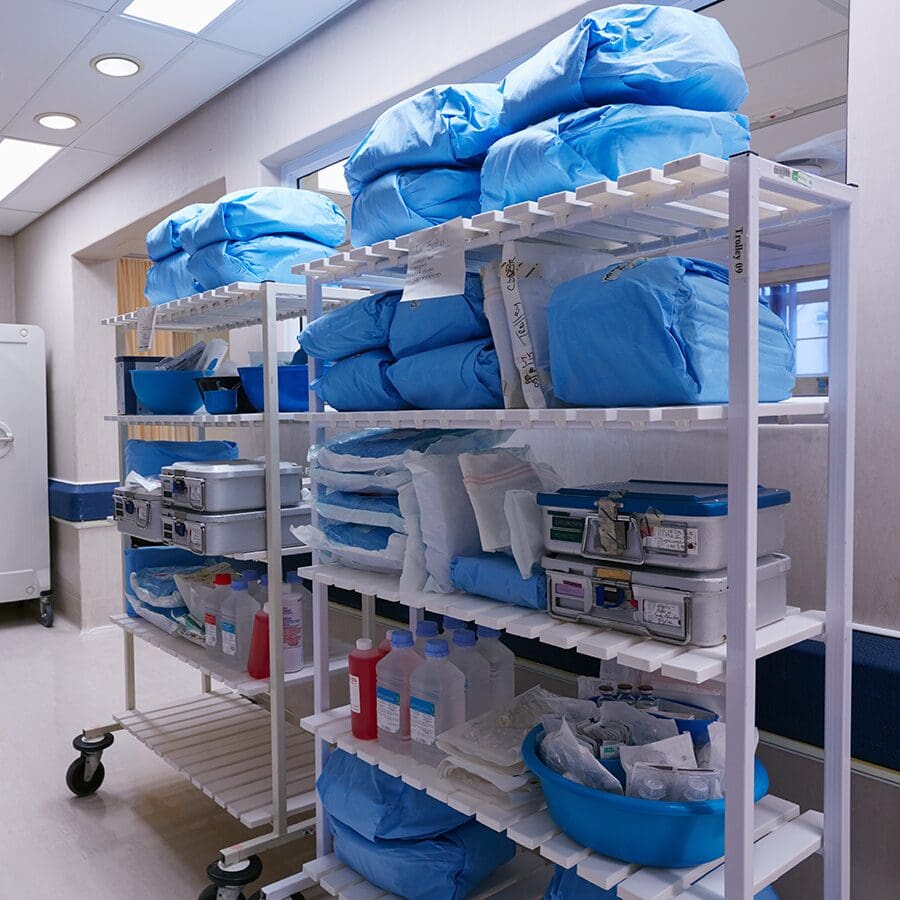 Two white racks in a hospital room hold blue-wrapped sterile supplies, blue trays with medical instruments, bottled liquids, and other medical equipment organized on multiple shelves.