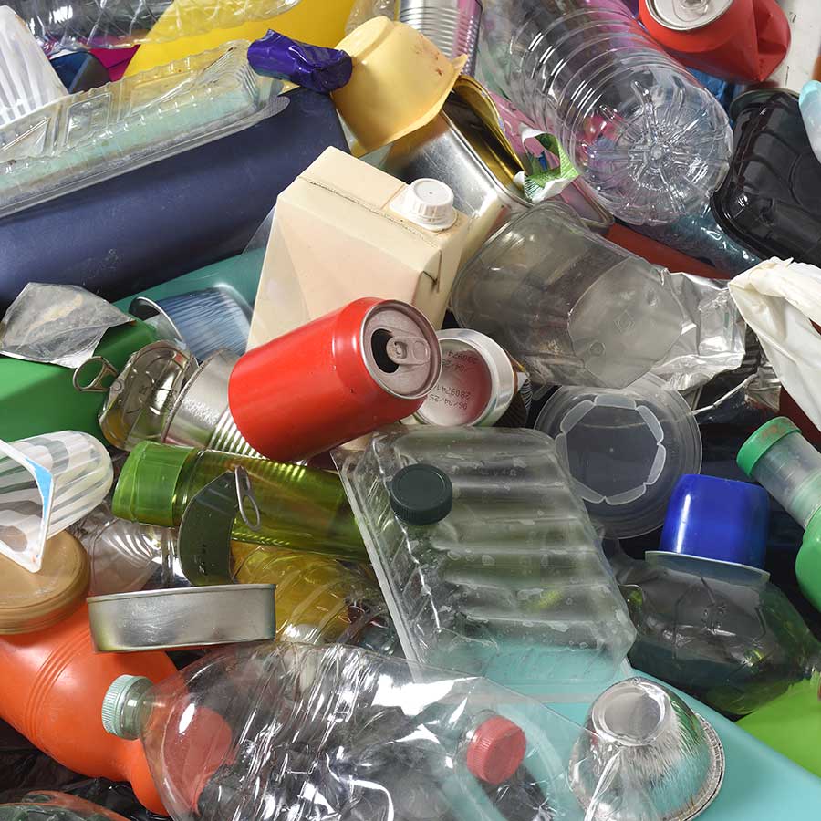 A pile of mixed recyclable waste, including plastic bottles, metal cans, food containers, cartons, and various packaging materials in different shapes and colors.