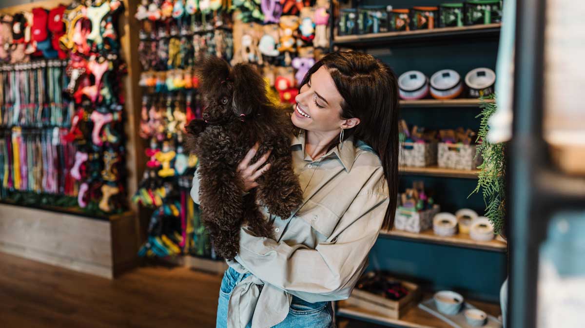 A woman smiles while holding a small, curly-haired brown dog inside a pet store, with colorful pet toys and supplies displayed on shelves in the background.
