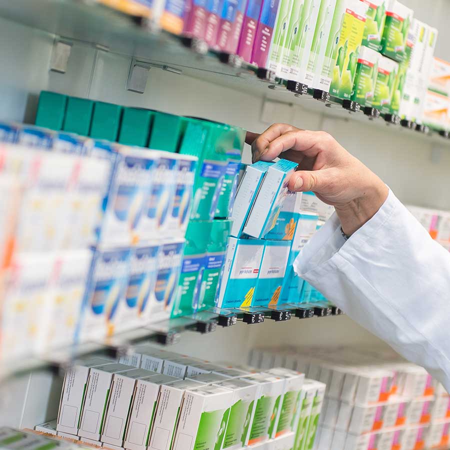 A person wearing a white coat selects a box of medicine from a shelf stocked with various pharmaceutical products in a pharmacy.