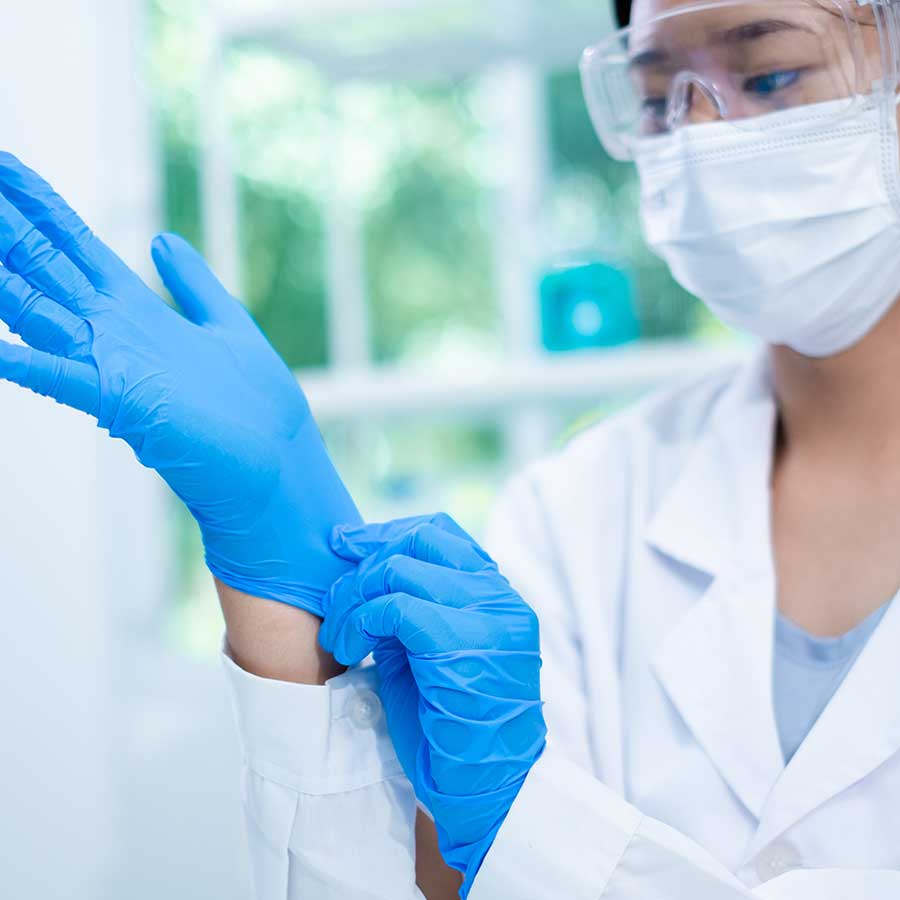 A person wearing a lab coat, safety glasses, and a face mask puts on blue disposable gloves in a bright, clinical setting.