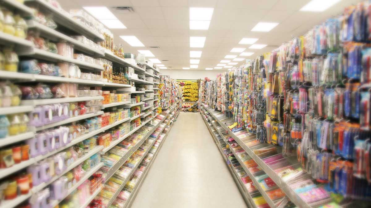 A brightly lit store aisle lined with shelves full of various colorful products, including household items, stationery, and packaged goods, extending to the back of the store.